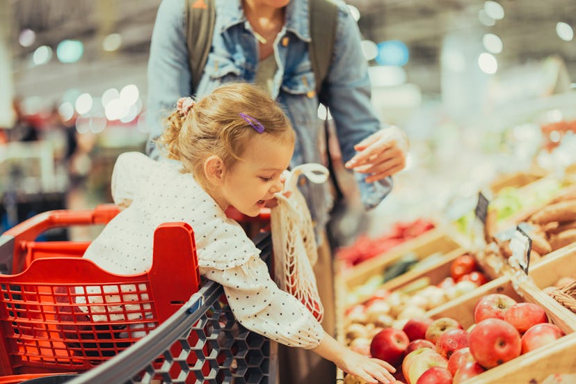 Close up shot of a cute cheerful girl sitting in the shopping cart and picking up apples in the frui...