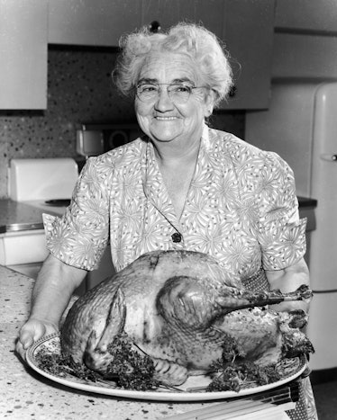 circa 1945:  An elderly woman in a dress and apron holds a large roasted turkey in a kitchen and smi...