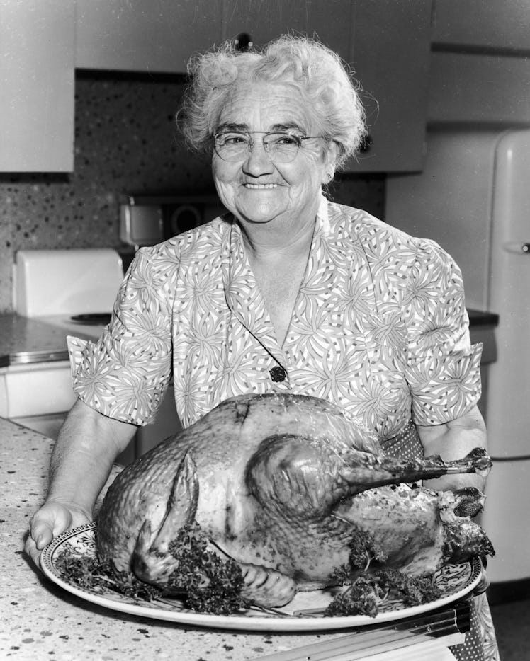 circa 1945:  An elderly woman in a dress and apron holds a large roasted turkey in a kitchen and smi...