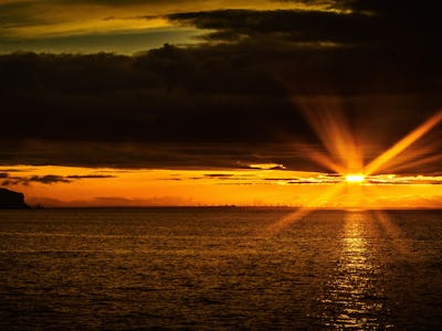 A coastal sunset with the landscape in shadow with a large glare from the sun as breaks through the ...