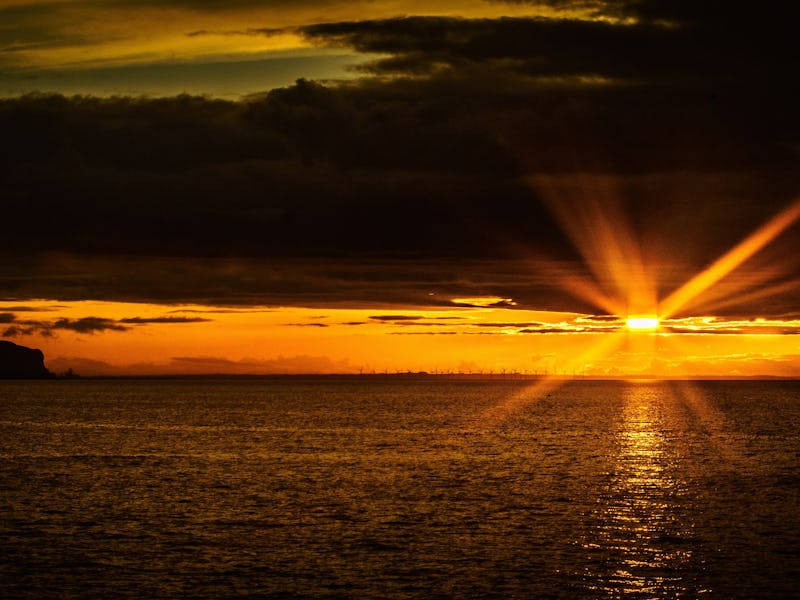 A coastal sunset with the landscape in shadow with a large glare from the sun as breaks through the ...