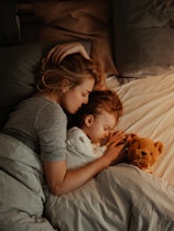Beautiful mother and daughter sleeping together in bed. It is the evening and there is lamp on. Mom ...