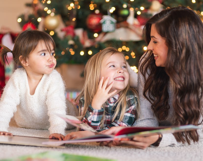 A young mom lies on the carpet by the twinkling lights on the Christmas tree and reads a book with h...