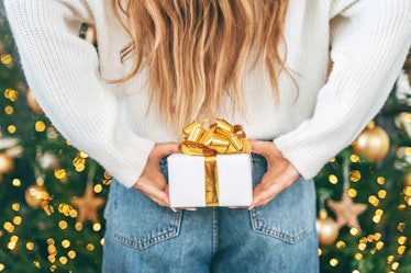 Woman in pastel white wool sweater hiding a small white gift box with golden ribbon bow behind her b...
