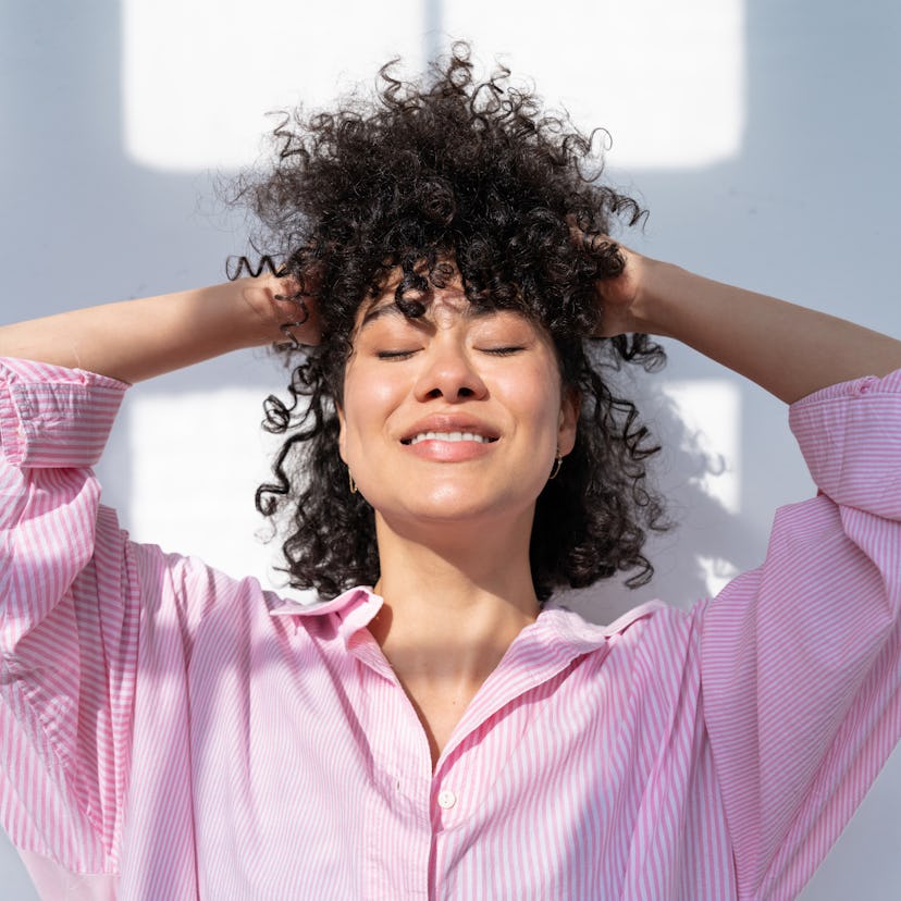 young woman closes her eyes and smiles as she adjusts her hair and considers the spiritual meaning o...