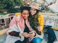 Two young women sit on a colorful public bench with their arms around each other, one holds up a cre...