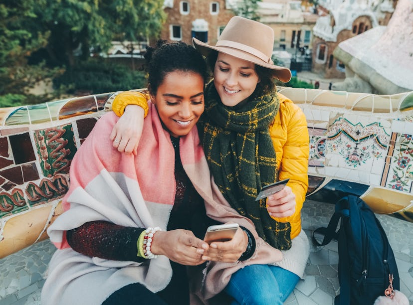 Two young women sit on a colorful public bench with their arms around each other, one holds up a cre...