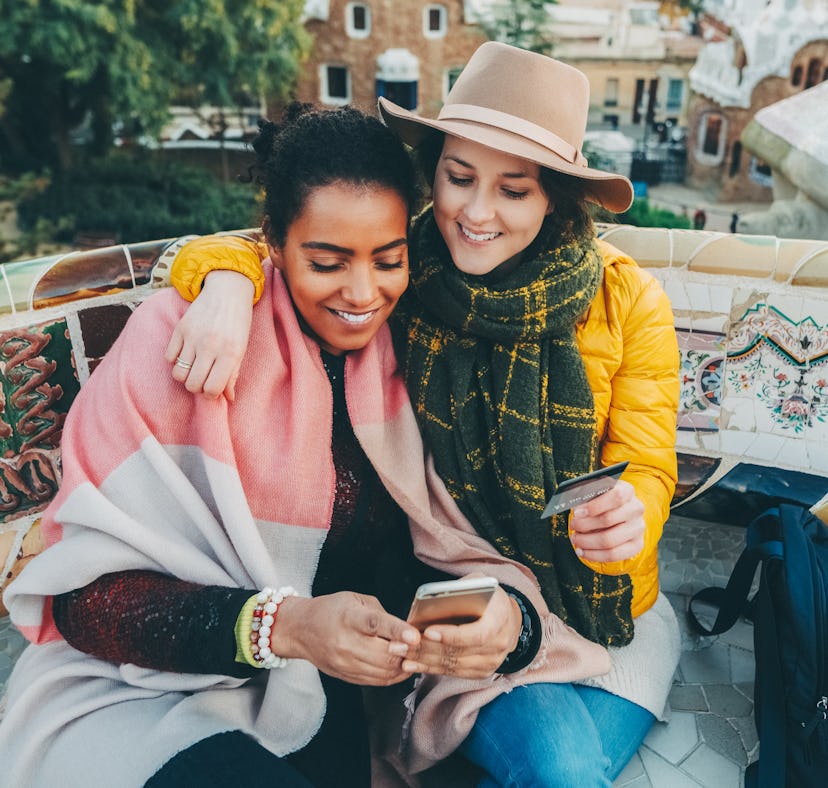 Two young women sit on a colorful public bench with their arms around each other, one holds up a cre...
