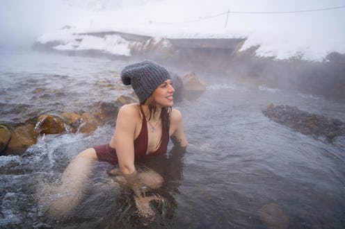 Young red-haired woman in burgundy swimsuit and gray cap is bathing in a thermal river in Iceland in...