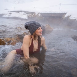 Young red-haired woman in burgundy swimsuit and gray cap is bathing in a thermal river in Iceland in...