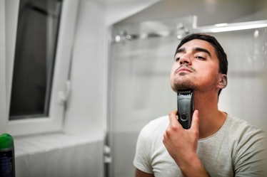 A young man is using an electric trimmer and shaving his beard in front of the mirror in a domestic ...