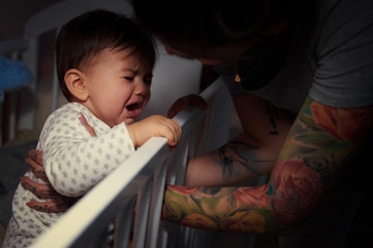 A parent comforts a crying toddler in a crib as they learn how to fall back and adjust a sleep sched...
