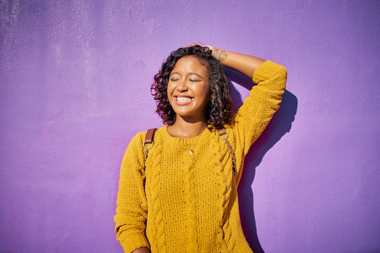 young woman poses in front of a purple wall and smiles, as she thinks about her november 14, 2022 we...