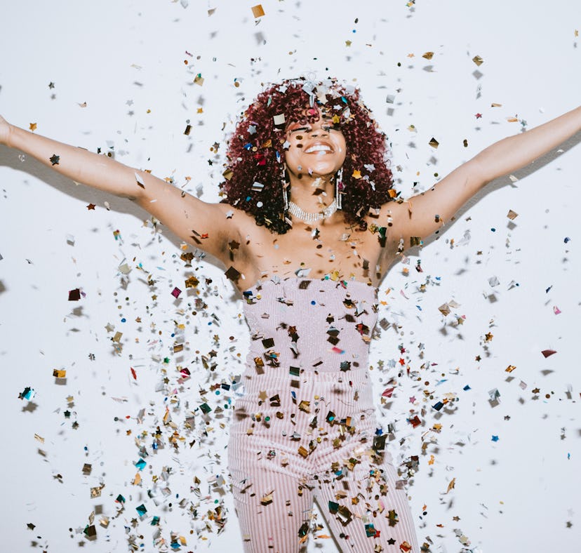 A young woman, dopamine dressed in a sparkling gown for the 2022 holidays, throws confetti in the ai...