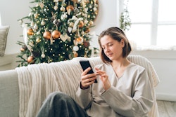 Young woman is using smartphone sitting on sofa near decorated Christmas tree with many lights in li...