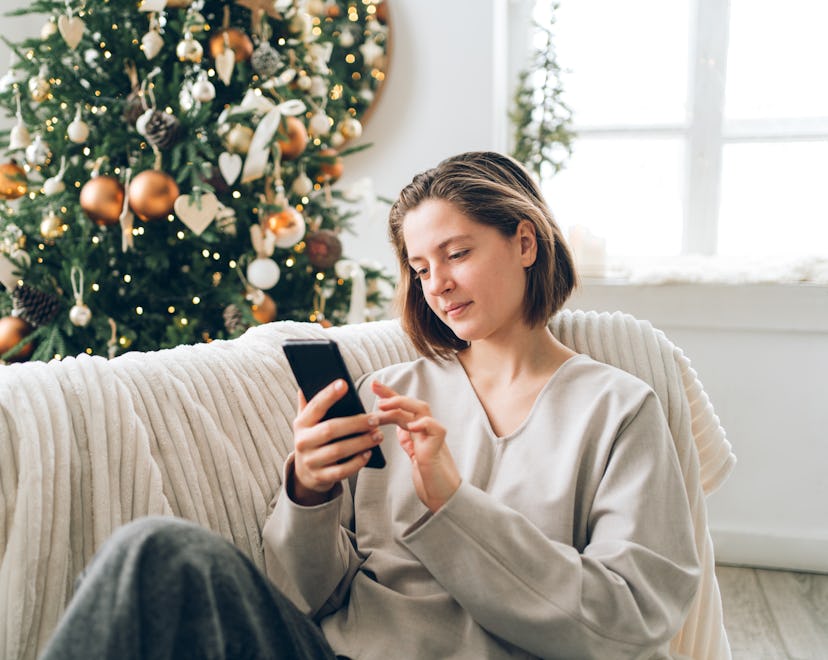 Young woman is using smartphone sitting on sofa near decorated Christmas tree with many lights in li...