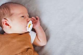 Newborn baby lying on bed and suckling a pacifier. Head shaping pillows are not safe for babies and ...