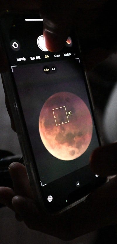 A man uses a smartphone to take picture of a blood moon through telescope during a total lunar eclip...