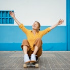 young woman sits on her skateboard and smiles with her arms open as she thinks about how the novembe...