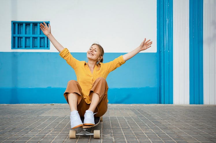 young woman sits on her skateboard and smiles with her arms open as she thinks about how the novembe...