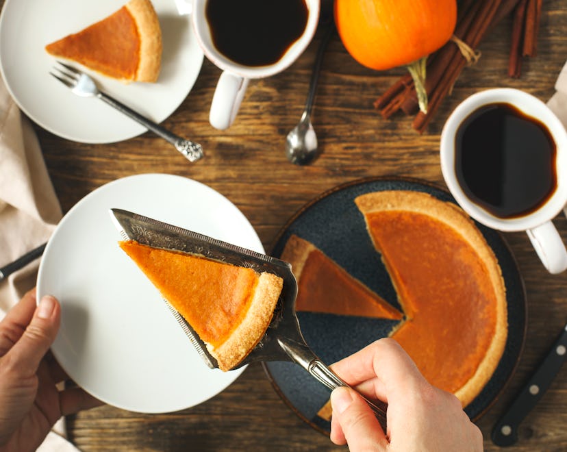A pumpkin pie on a table with coffee mugs and hands serving a slice of pie on a plate in the foregro...