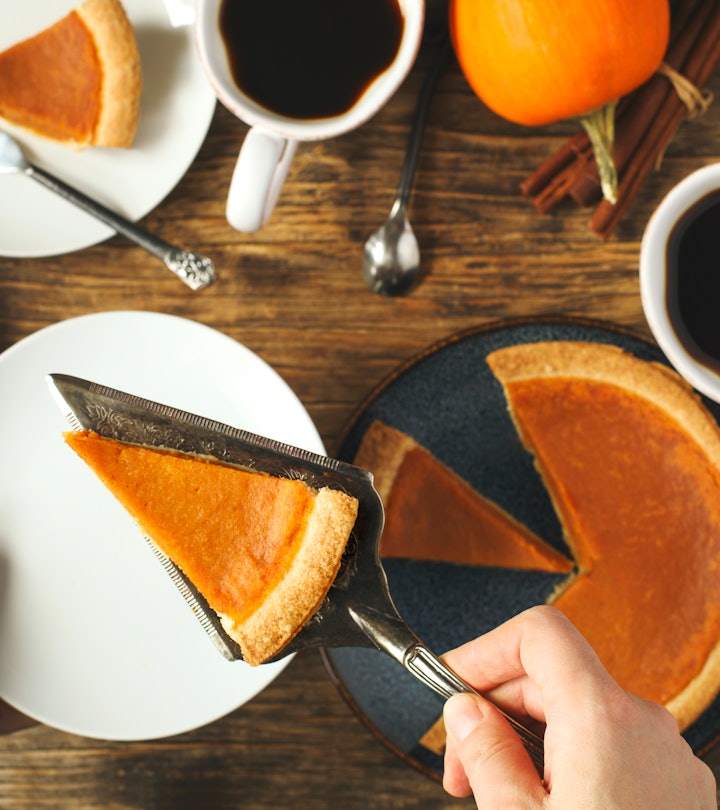 A pumpkin pie on a table with coffee mugs and hands serving a slice of pie on a plate in the foregro...
