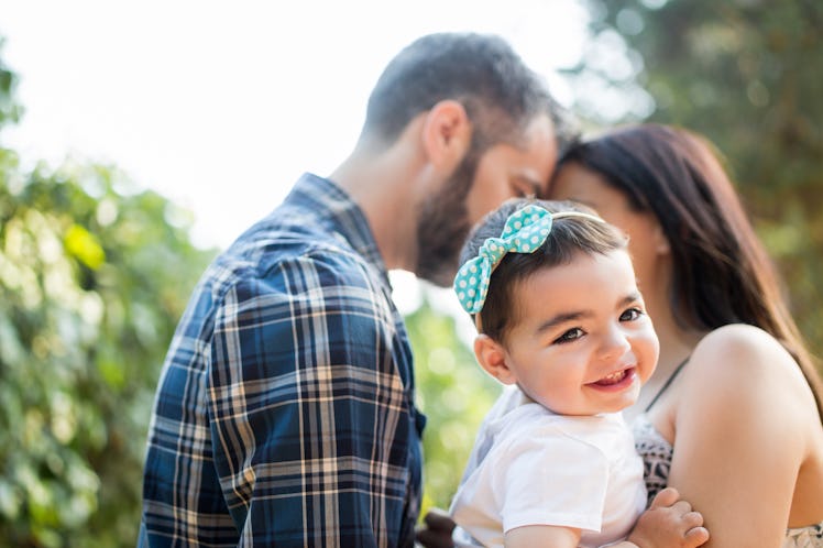 A happy latin baby girl in her parents' arms smiling away in a horizontal waist up shot outdoors.
