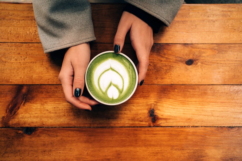 Woman hands hold a ceramic cup with matcha green tea latte with foam art on the wooden table. Backdr...