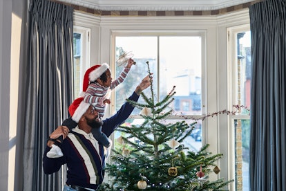 Daughter sits on father's shoulders to put a star on top of a Christmas tree in bay window