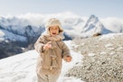 甜美的宝宝穿着时尚的冬装，背景是雪山。孩子穿着温暖的夹克…
