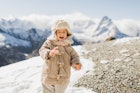 Sweet baby in stylish winter clothing on background of snowy mountains. Child wearing in warm jacket...