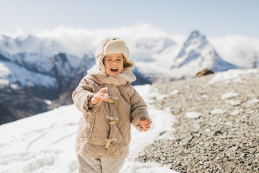 Sweet baby in stylish winter clothing on background of snowy mountains. Child wearing in warm jacket...