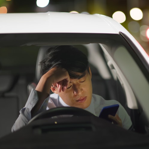 担心的亚洲商人晚上开车上班时用智能手机工作，他觉得很累