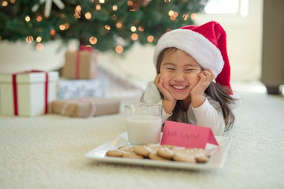 Adorable young girl wearing Santa hat lays in front of a Christmas tree with a tray of cookies and g...