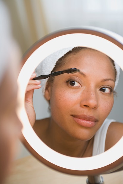 A Black woman grooming her eyebrows in front of a mirror. 