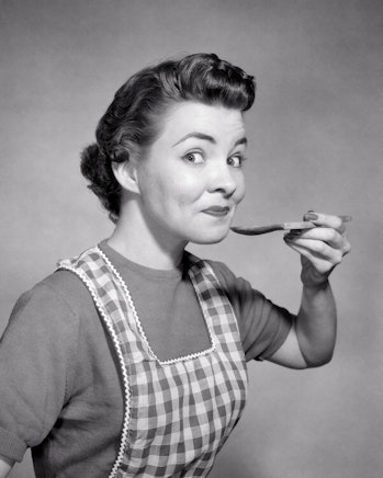 1950s smiling woman housewife wearing checkered apron looking at camera with raised eyebrows tasting...