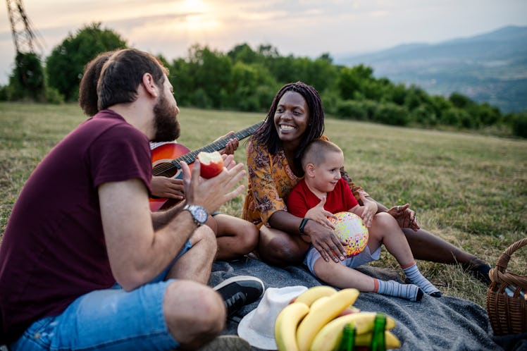 Happy diverse family with two children sitting on picnic blanket, playing guitar and singing