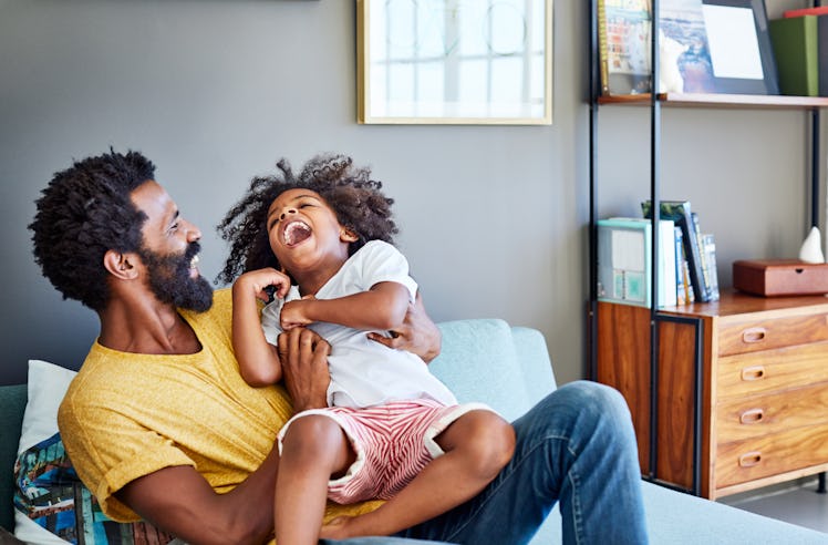Shot of a cheerful young man holding his son and ticking him while being seated on a couch at home