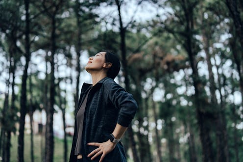 Relaxed young Asian sports woman with eyes closed breathing fresh air in nature park with trees in b...