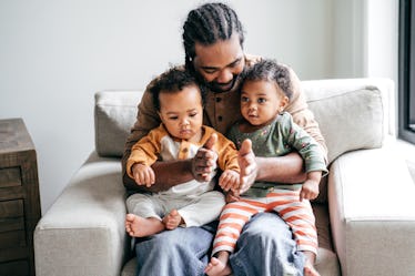 A dad on a chair smiling down at his twin daughters in his lap.