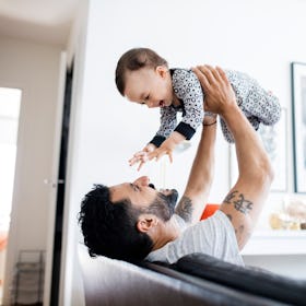 A father sitting on a couch holds his baby in the air.