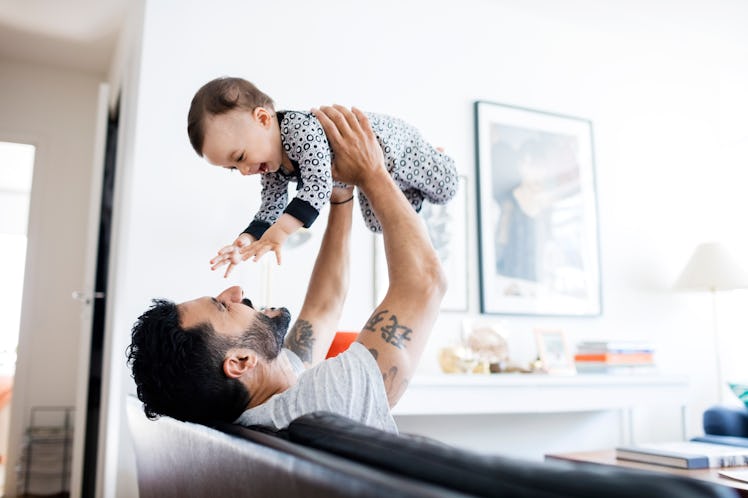 A father sitting on a couch holds his baby in the air.