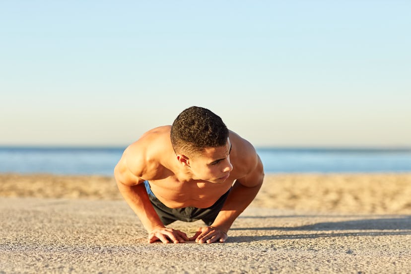 Shirtless young male athlete doing diamond push-ups at beach. Muscular man exercising against sky. H...