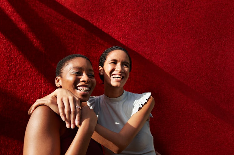 Smiling young woman standing with female friend against red wall thinking about their luckiest day i...