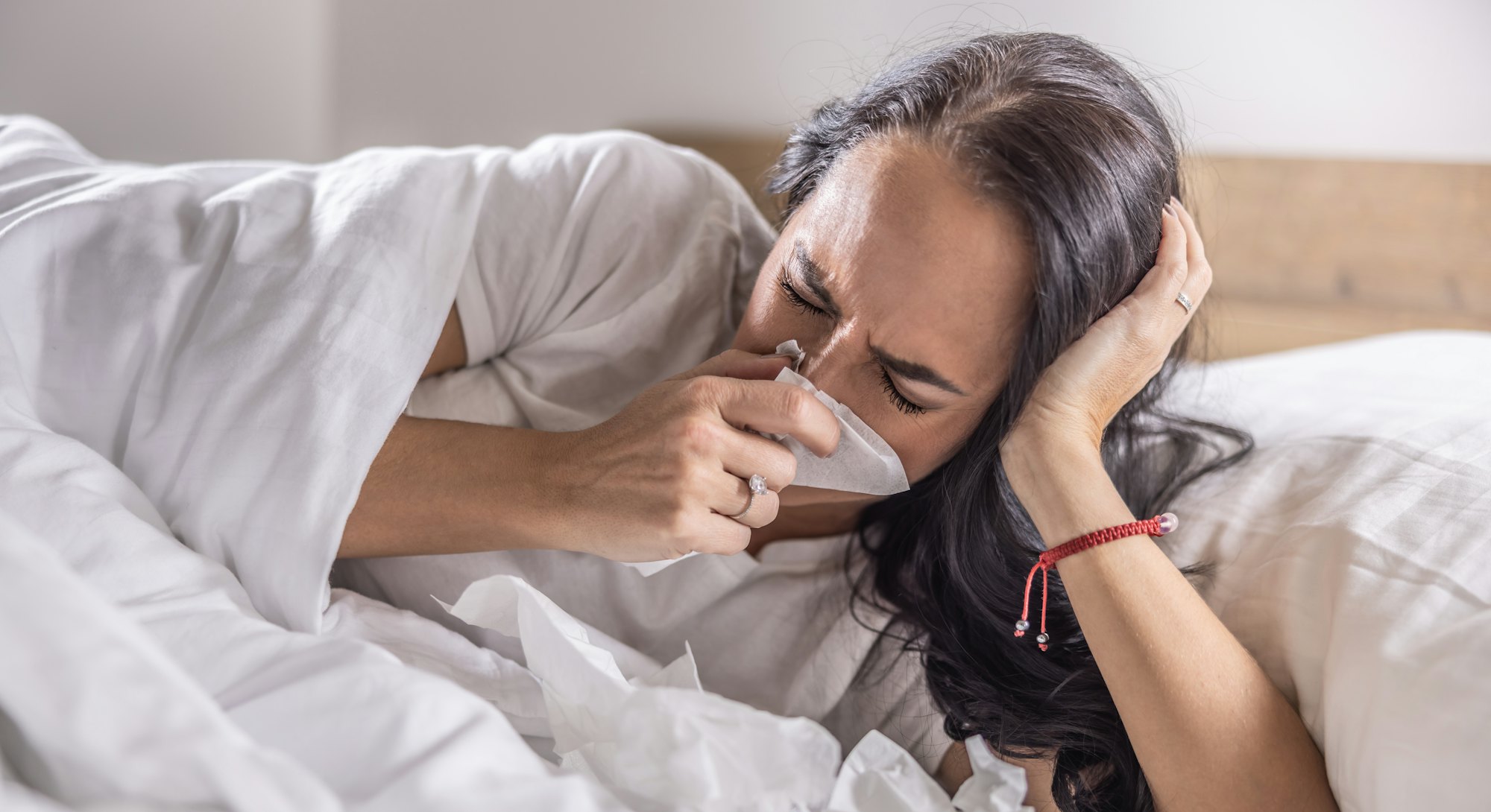 A sick woman sneezes into a napkin, lies in bed, has the flu or a cold.