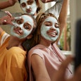 Happy young woman taking selfie with female friends wearing facial cream in bathroom at home
