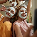 Happy young woman taking selfie with female friends wearing facial cream in bathroom at home