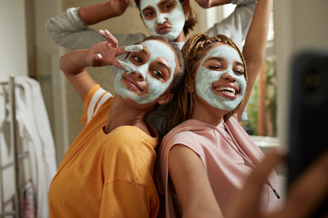 Happy young woman taking selfie with female friends wearing facial cream in bathroom at home