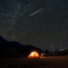 A shooting star in the sky above a person camping out in a tent near mountains.
