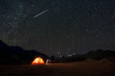 A shooting star in the sky above a person camping out in a tent near mountains.